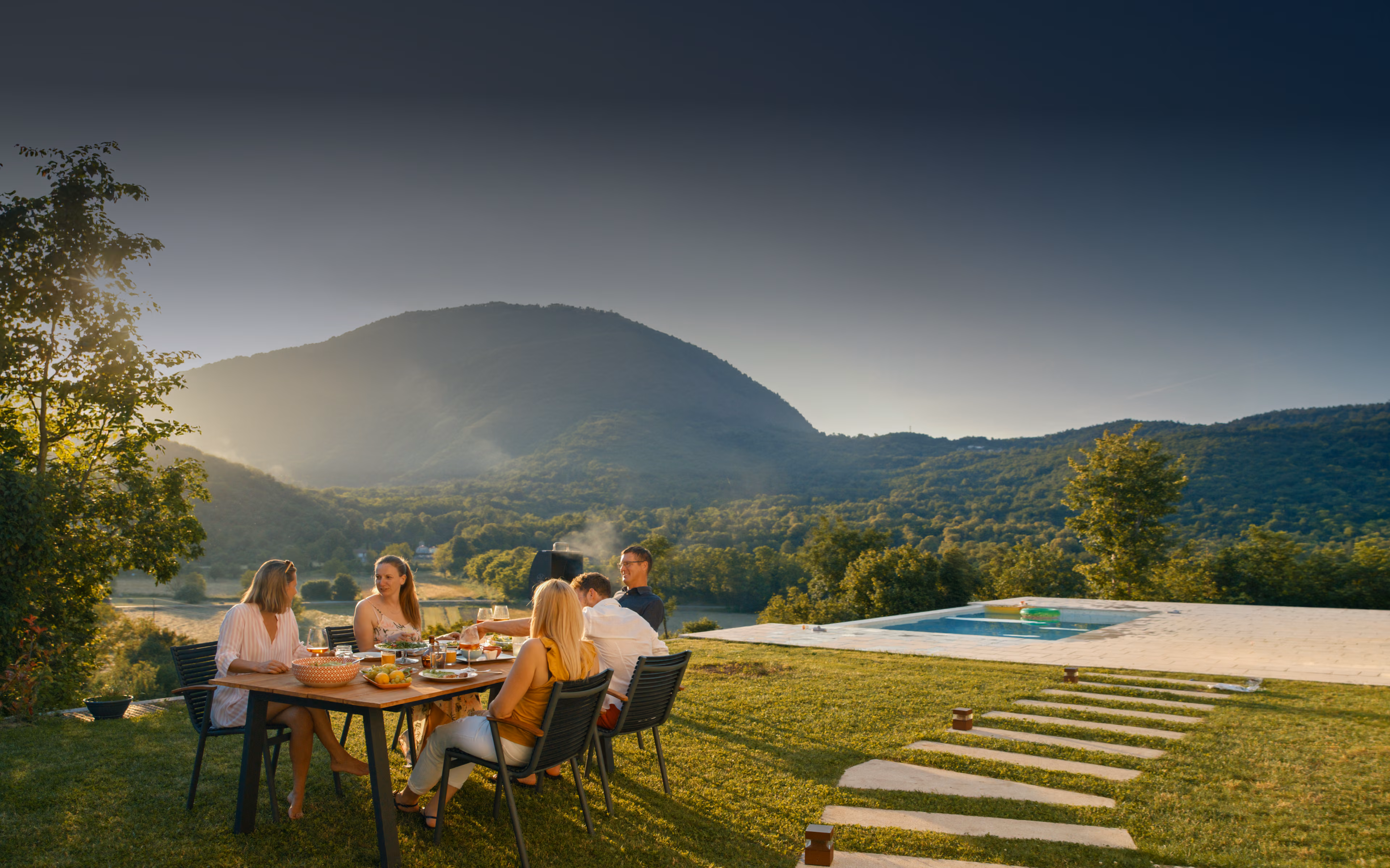 Family dining outdoors on a sunlit hillside with mountain views and pool.