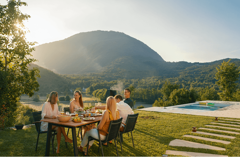 Family dining outdoors on a sunlit hillside with mountain views and pool.