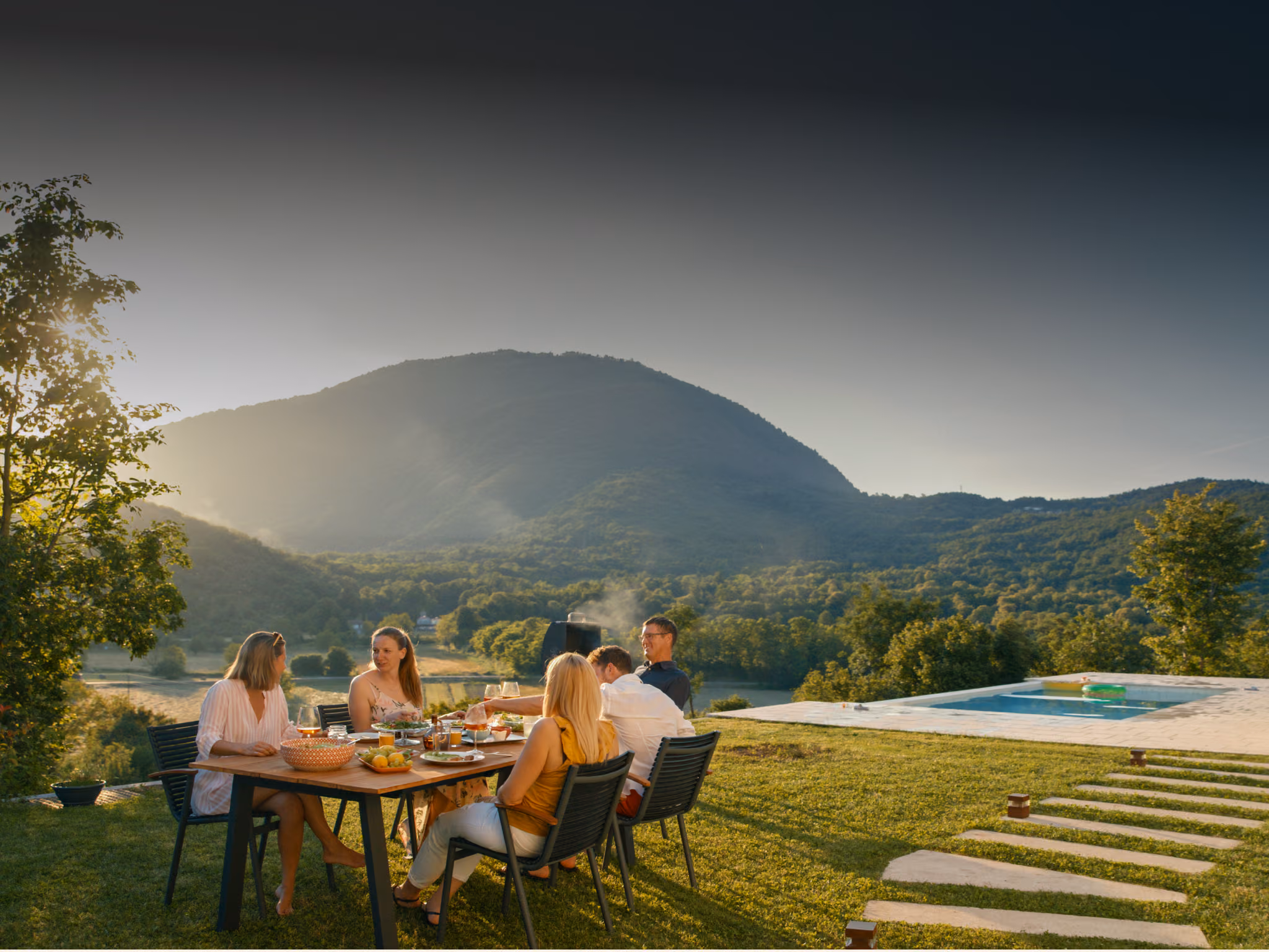 Family dining outdoors on a sunlit hillside with mountain views and pool.